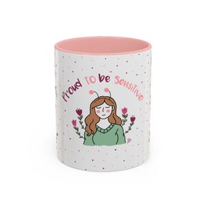 Könnte beinhalten: Weißer Keramikbecher mit rosa Innenraum. Der Becher zeigt den Text "proud to be sensitive" und eine Cartoon-Illustration einer Frau mit braunen Haaren, grünem Pullover und floralen Akzenten.