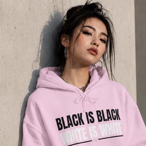 Puede incluir: Una sudadera con capucha de color rosa claro que presenta un gráfico frontal con el texto BLACK IS BLACK en letras negras en negrita y WHITE IS WHITE en letras blancas, separado por una fina línea horizontal negra y un bolsillo frontal.