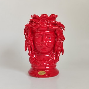 May include: A vibrant red ceramic vase shaped like a human head with a mustache and a crown of chili peppers. The vase has a glossy finish and a round base. The base has a small, round sticker with text.
