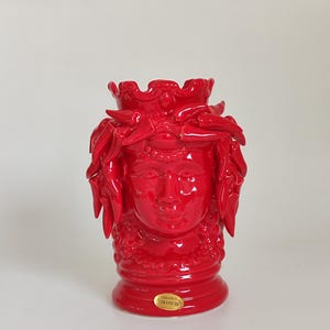 May include: A vibrant red ceramic vase shaped like a classical female head with ornate details. The vase features a crown-like top and a detailed face. A small, round sticker with text is visible on the base. The vase is a decorative item.