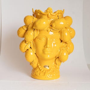 Könnte beinhalten: Eine gelbe Keramikvase in Form eines Frauenkopfes mit Zitronen um ihren Kopf. Die Vase ist mit einer Krone und einer Halskette verziert.