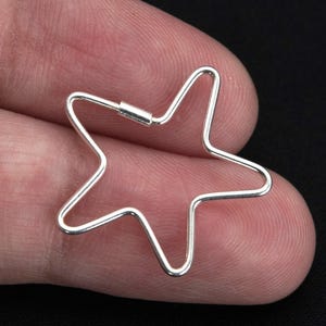 May include: A silver-coloured star-shaped charm. The charm is made of thin metal wire bent into a five-pointed star. The charm is held in a person's hand against a black background.