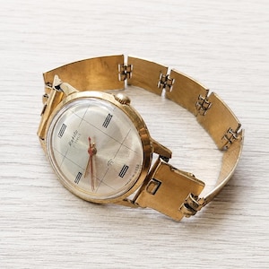 Op de afbeelding: Een goudkleurig Raketa-polshorloge met een zilveren wijzerplaat en zwarte markeringen. Het horloge heeft een goudkleurige band met een sluiting. De wijzerplaat heeft de tekst "Raketa" en "23 Jewels". Het horloge is gemaakt in de USSR.