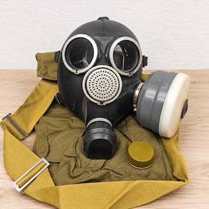 Op de afbeelding: Een zwart rubberen gasmasker met twee ronde glazen ooglenzen met zilveren randen, een centrale ronde geperforeerde metalen filterbehuizing en een aan de zijkant gemonteerde grijs-witte cilindrische filterbus, vergezeld van een olijfgroene canvas tas.
