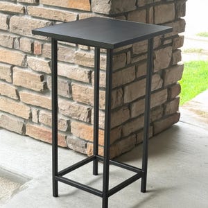 May include: A black metal side table with a square top and a geometric frame. The table has a height of approximately 30 inches and is suitable for indoor or outdoor use. The table is made of metal and has a minimalist design.