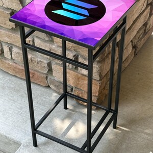 K&ouml;nnte beinhalten: Ein schwarzer Beistelltisch aus Metall mit quadratischer Platte. Die Tischplatte hat ein geometrisches Design in Lila- und Blaut&ouml;nen mit einem kreisf&ouml;rmigen Logo in der Mitte. Der Tisch ist etwa 61 cm hoch.