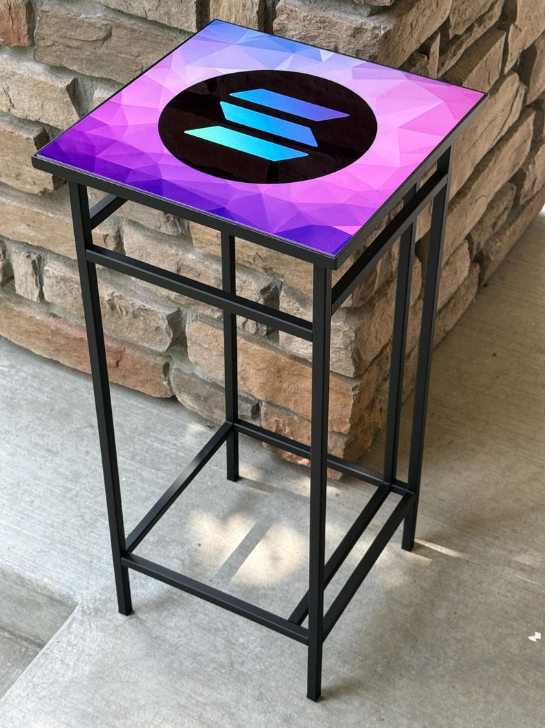 K&ouml;nnte beinhalten: Ein schwarzer Beistelltisch aus Metall mit quadratischer Platte. Die Tischplatte hat ein geometrisches Design in Lila- und Blaut&ouml;nen mit einem kreisf&ouml;rmigen Logo in der Mitte. Der Tisch ist etwa 61 cm hoch.
