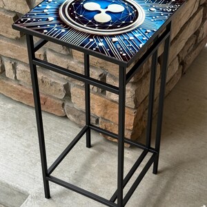 K&ouml;nnte beinhalten: Ein schwarzer Metall-Beistelltisch mit quadratischer Platte mit blauem und wei&szlig;em Design. Das Design umfasst ein Leiterplattenmuster und ein zentrales Ripple-Logo. Der Tisch ist etwa 61 cm hoch.