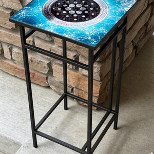 K&ouml;nnte beinhalten: Ein quadratischer Beistelltisch mit schwarzem Metallrahmen. Die Tischplatte hat ein blau-wei&szlig;es Design mit einer zentralen kreisf&ouml;rmigen Grafik, die einem Kryptow&auml;hrungssymbol &auml;hnelt. Der Tisch ist etwa 61 cm hoch.