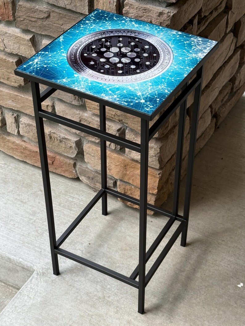 K&ouml;nnte beinhalten: Ein quadratischer Beistelltisch mit schwarzem Metallrahmen. Die Tischplatte hat ein blau-wei&szlig;es Design mit einer zentralen kreisf&ouml;rmigen Grafik, die einem Kryptow&auml;hrungssymbol &auml;hnelt. Der Tisch ist etwa 61 cm hoch.