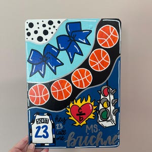 Puede incluir: Letrero rectangular pintado a mano con temática de baloncesto. Presenta balones de baloncesto, lazos azules y una camiseta con el nombre "SCOTT" y el número "23". Incluye el texto "hey 23, call me" y "MS. brichne".
