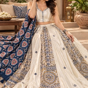 May include: A cream-colored sleeveless blouse with a V-neckline and a matching floor-length skirt featuring intricate blue, gold, and white patterned panels. A separate dark blue shawl with a repeating heart and sun motif is draped over one side.