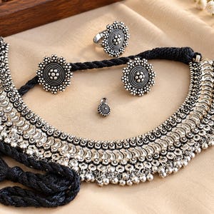 May include: A silver-toned jewelry set featuring a wide, crescent-shaped necklace with intricate metalwork and small dangling beads, paired with matching circular stud earrings, a circular ring, and a small circular pendant, all on black cord.