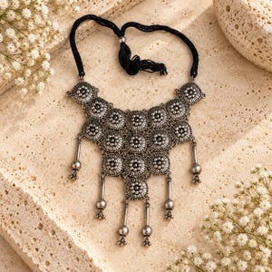 May include: A silver-toned metal statement necklace featuring a tiered arrangement of circular, floral-patterned medallions. The piece includes dangling cylindrical and spherical charms at the base and is secured by a black cord with a knotted tie.