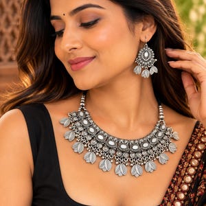 May include: A silver-toned metal necklace and matching drop earrings set featuring intricate filigree patterns, small white oval cabochons, and dangling leaf-shaped charms. The necklace has a wide, curved design that sits at the base of the neck.