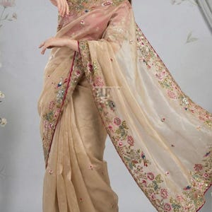May include: A beige saree with intricate floral embroidery and a contrasting magenta blouse. The saree features detailed embellishments along the borders and sleeves, creating an elegant and festive look. The fabric appears sheer and flowing, suitable for special occasions.