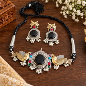 May include: A jewelry set featuring a black cord necklace and matching earrings. The necklace has a central silver-toned pendant with a black stone, flanked by gold-toned bird figures, colorful rectangular stones, and small white bead dangles.
