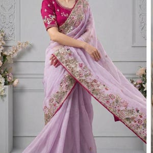 May include: A light purple saree with intricate embroidery along the borders and a matching magenta blouse. The saree features floral and peacock motifs with gold accents. The fabric appears lightweight and flowing, suitable for special occasions.