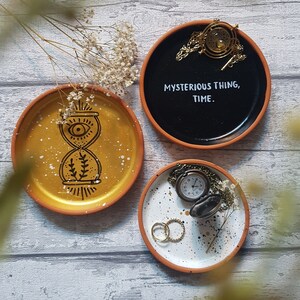 Könnte beinhalten: Drei dekorative Keramik-Schmuckschalen mit goldenen und weißen Akzenten. Eine Schale hat einen schwarzen Hintergrund mit dem Text "MYSTERIOUS THING, TIME". Eine andere Schale hat einen goldenen Hintergrund mit einem weißen Sanduhr-Design. Die dritte Schale hat einen weißen Hintergrund mit schwarzen Flecken und einer goldenen Taschenuhr.