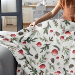 Puede incluir: Una manta blanca con un patrón navideño festivo con adornos rojos, piñas y ramas de pino verdes. La manta se coloca sobre un sofá gris. El tema general es la decoración navideña.