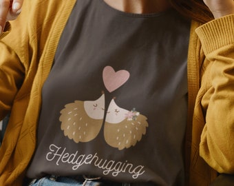 Könnte beinhalten: Ein dunkelgraues T-Shirt mit einem Motiv von zwei sich umarmenden Igeln mit einem rosa Herzen darüber. Der Text "Hedgehugging" ist unter den Igeln gedruckt.