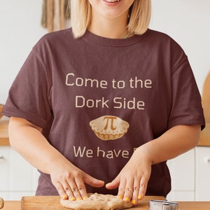 May include: A person wearing a maroon t-shirt with the text "Come to the Dork Side" and a pie with the symbol for pi on it. The text "We have pi" is below the pie. The person is making a pie crust.
