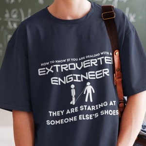 May include: A navy blue t-shirt with white text that reads "HOW TO KNOW IF YOU ARE DEALING WITH AN EXTROVERTED ENGINEER THEY ARE STARING AT SOMEONE ELSE'S SHOES". The text is accompanied by a simple stick figure illustration.