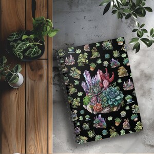 Puede incluir: Una libreta espiral negra con una ilustración colorida de suculentas y cristales. La portada presenta un patrón repetitivo de pequeñas suculentas y cristales sobre un fondo negro.