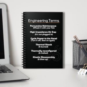 May include: A black spiral bound notebook with the title "Engineering Terms" and humorous definitions of technical terms like "Percussive Maintenance" and "Thermal Shock".