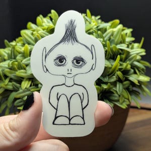 May include: A die-cut sticker featuring a minimalist illustration of a seated figure with a sad expression. The black line art depicts a person with large eyes, a small mouth, and messy hair. The sticker is held against a background of green foliage.
