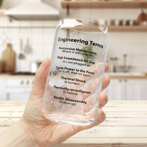 May include: A clear glass can with a white label that reads "Engineering Terms" and lists humorous definitions for common engineering terms. The can is being held by a hand.