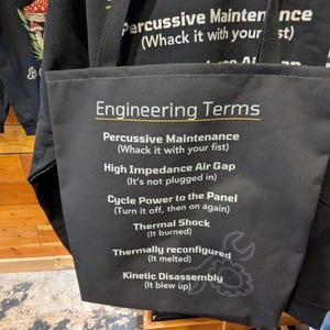 May include: Black tote bag featuring the words "Engineering Terms" in white. The bag lists humorous engineering terms such as "Percussive Maintenance" and "Thermal Shock". A black strap is visible.