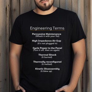 May include: Black t-shirt with the text "Engineering Terms" and humorous definitions, including "Percussive Maintenance" and "Kinetic Disassembly." The shirt is worn by a person with a beard.