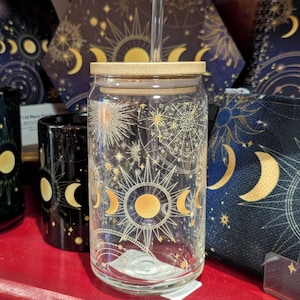 May include: A clear glass tumbler with a bamboo lid and a glass straw, featuring a gold and white celestial print of suns, moons, and stars. The design includes intricate geometric patterns and phases of the moon encircling the cylindrical glass.