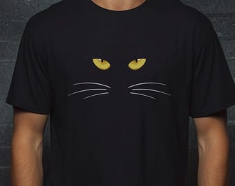 May include: A black short-sleeved t-shirt featuring a graphic print of two piercing yellow cat eyes positioned above three thin white curved lines representing whiskers, centered on the chest area of the garment.