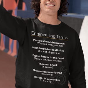May include: A black sweatshirt with a white graphic print that defines common engineering terms in a humorous way. The text includes "Percussive Maintenance (Whack it with your fist)", "High Impedance Air Gap (It's not plugged in)", "Cycle Power to the Panel (Turn it off, then on again)", "Thermal Shock (It burned)", "Thermally reconfigured (It melted)", and "Kinetic Disassembly (It blew up)".