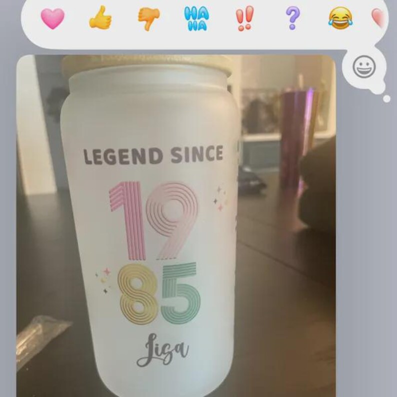 May include: A frosted glass jar with a wooden lid. The jar is decorated with the text "LEGEND SINCE" above the numbers "1985" in colorful fonts, and the name "Jiga" below. The jar is on a dark surface.