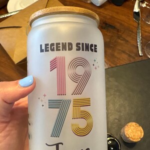 May include: Frosted glass jar with a wooden lid, featuring the text "LEGEND SINCE 1975 Janae" in colorful lettering. The jar is held by a person with light blue nail polish. A cork and a black bottle stopper are on the table.