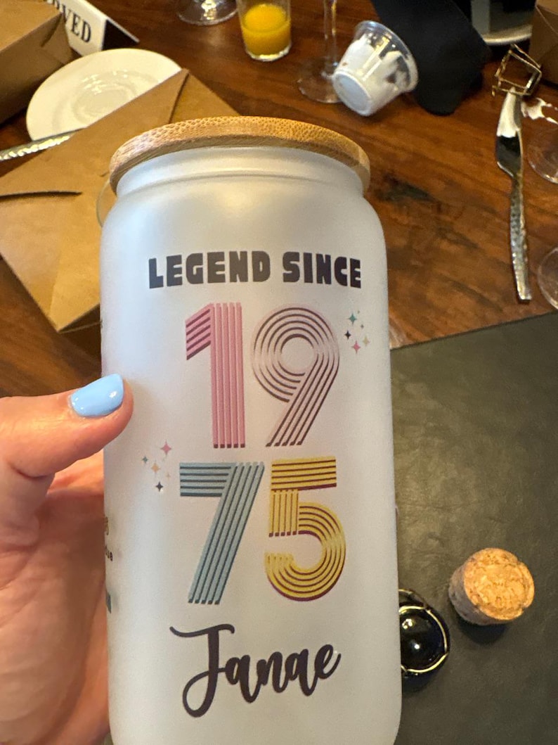 May include: Frosted glass jar with a wooden lid, featuring the text "LEGEND SINCE 1975 Janae" in colorful lettering. The jar is held by a person with light blue nail polish. A cork and a black bottle stopper are on the table.