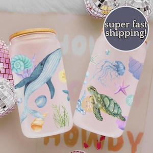May include: Two glass tumblers with bamboo lids. One features a blue whale, the other a sea turtle, with seashell and starfish designs. The tumblers have a pink background and are decorated with watercolor illustrations.