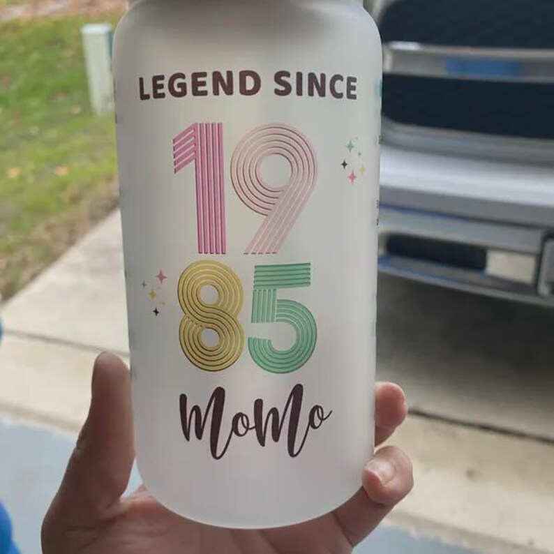 May include: A white water bottle with the text "LEGEND SINCE 1985 Momo" in pink, gold, and green. The bottle has a frosted appearance and is held by a person.