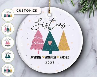 May include: A round white ceramic ornament featuring the word Sisters in black script, three stylized Christmas trees in pink, teal, and gold, and the names Jasmine, Amanda, and Harper separated by gold hearts with the year 2027 below.