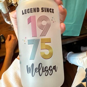 May include: Frosted glass jar with a bamboo lid. The jar is decorated with the text "LEGEND SINCE 1975 Melissa" in a colorful design. The numbers are pink, blue, and yellow. The jar is held by a person.