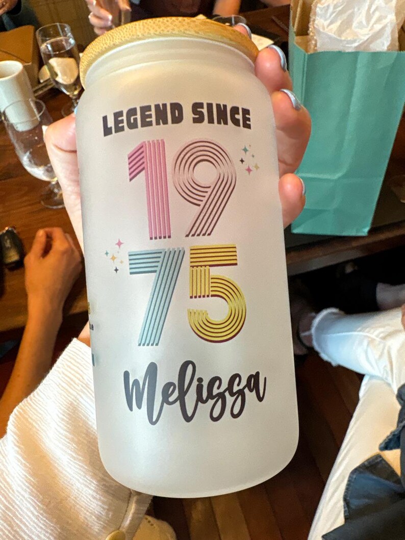 May include: Frosted glass jar with a bamboo lid. The jar is decorated with the text "LEGEND SINCE 1975 Melissa" in a colorful design. The numbers are pink, blue, and yellow. The jar is held by a person.