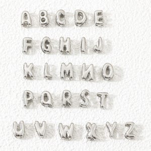 Op de afbeelding: Een verzameling van zesentwintig individuele zilverkleurige metalen alfabetletterkralen, gerangschikt in vijf rijen van A tot Z. Elke letter heeft een glad, afgerond en bol ontwerp met een reflecterende metalen afwerking voor sieraden.