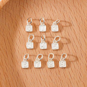 May include: A collection of silver-colored number charms, each with a small loop for attachment. The numbers 0 through 9 are displayed on square, white-filled backgrounds. The charms are arranged on a light brown wooden surface.