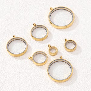 May include: A collection of six circular gold-toned metal floating locket pendants of varying sizes. Each pendant features a clear center, a thin metallic rim, and a small loop at the top for attachment, arranged in a scattered pattern.