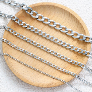 May include: An assortment of silver-colored chain necklaces of varying thicknesses, displayed on a light brown wooden plate. The chains feature different link styles, from thick curb chains to delicate, thin chains. The chains are arranged diagonally across the plate.