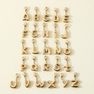 May include: A collection of gold-colored alphabet charms, each with a lobster clasp for easy attachment. The letters are in a rounded, bubble font, and are arranged in rows and columns against a neutral background.
