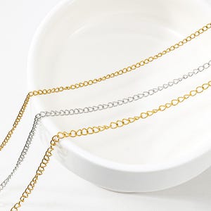 May include: Three delicate metal chains arranged in parallel lines. Two of the chains are gold-toned, and one central chain is silver-toned. Each chain features a series of small, interlocking oval links that create a continuous, flexible strand.